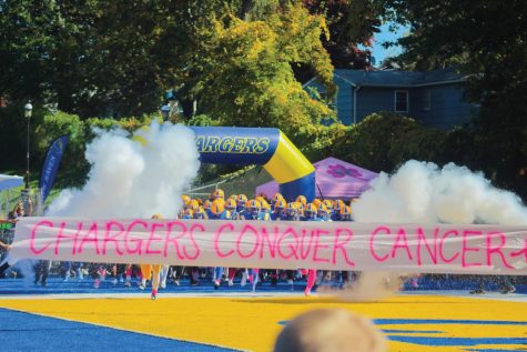 The New Haven football team runs through a Chargers Conquer Cancer banner before the game against Post, West Haven, Oct. 15, 2022. 